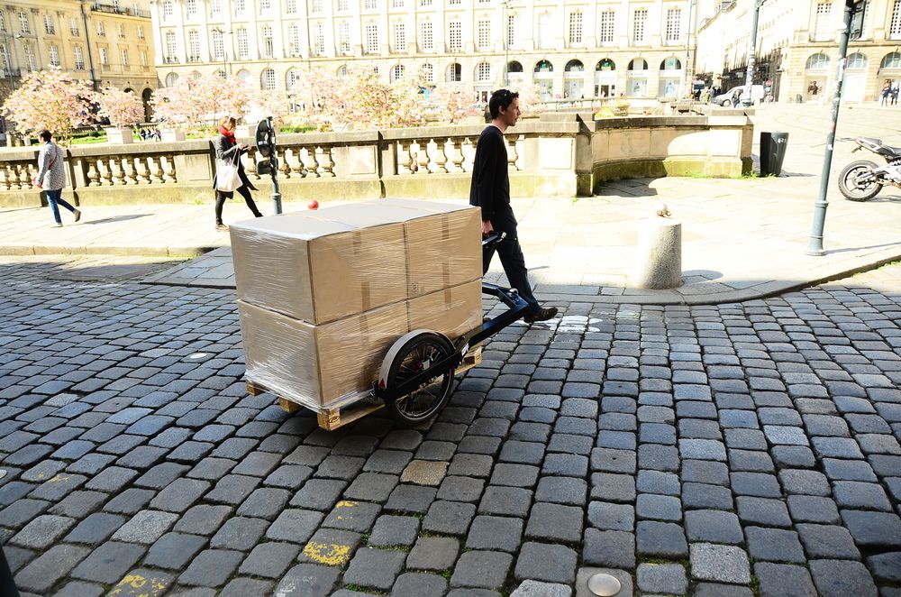 BicyLift bike trailer with 4 boxes on it being towed by hand on a paved street in an urban environment 