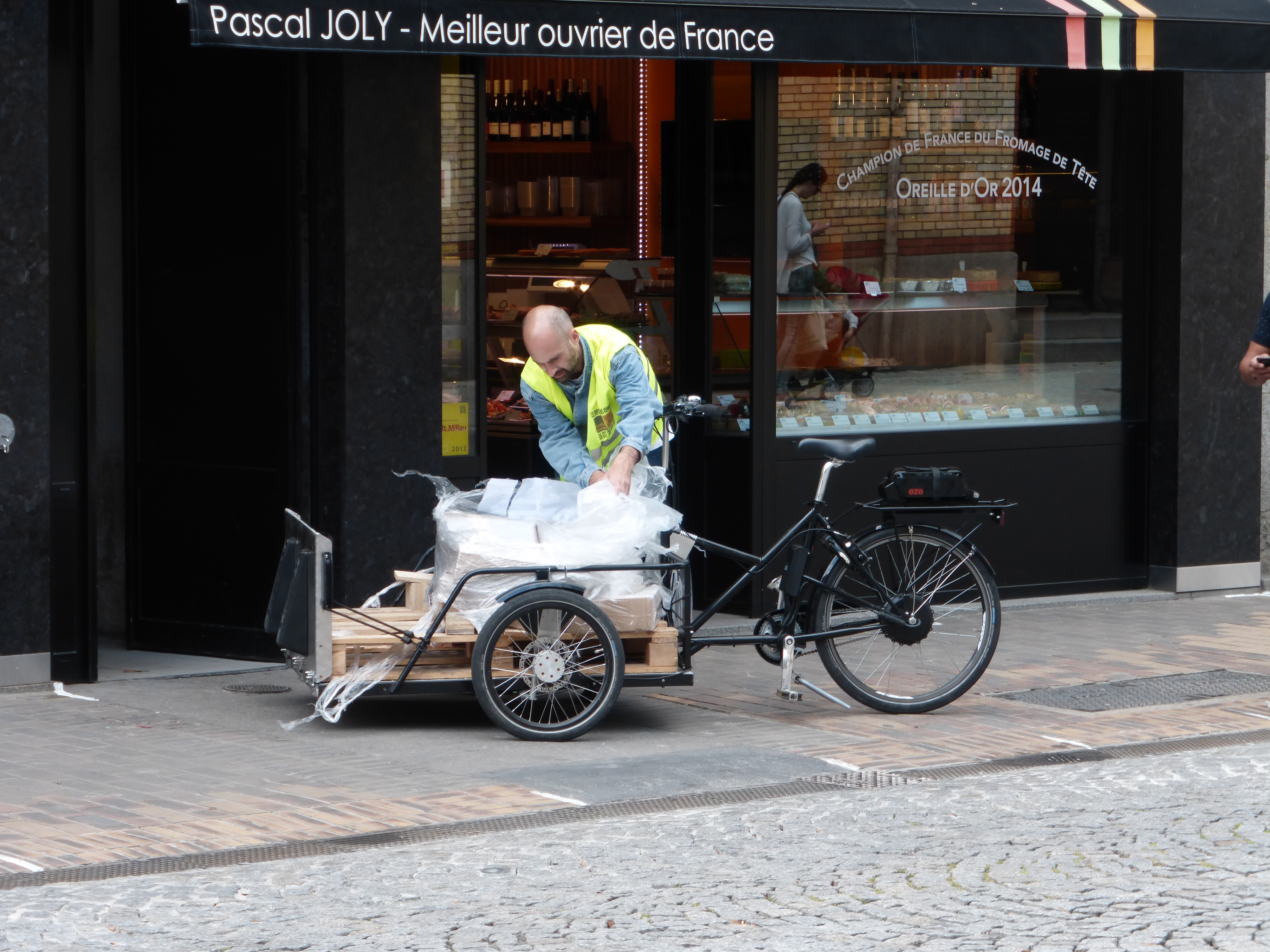 A man wearing a yellow jacket is unloading a cargo bike parked on the sidewalk, street is made of cobbles 