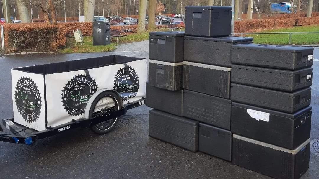 Isothermal boxes next to a bike trailer on which a big volume with white and black branding 