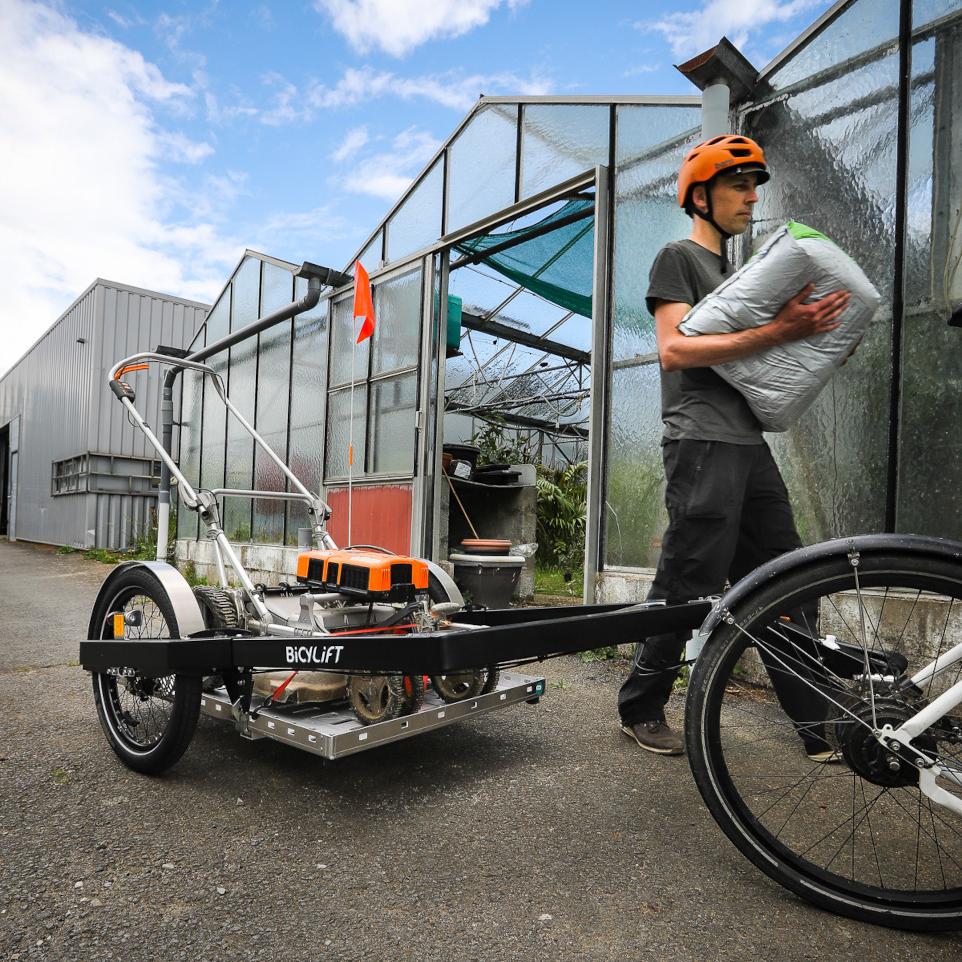 Person transporting compost in front of a bicycle trailer that carries a lawnmower