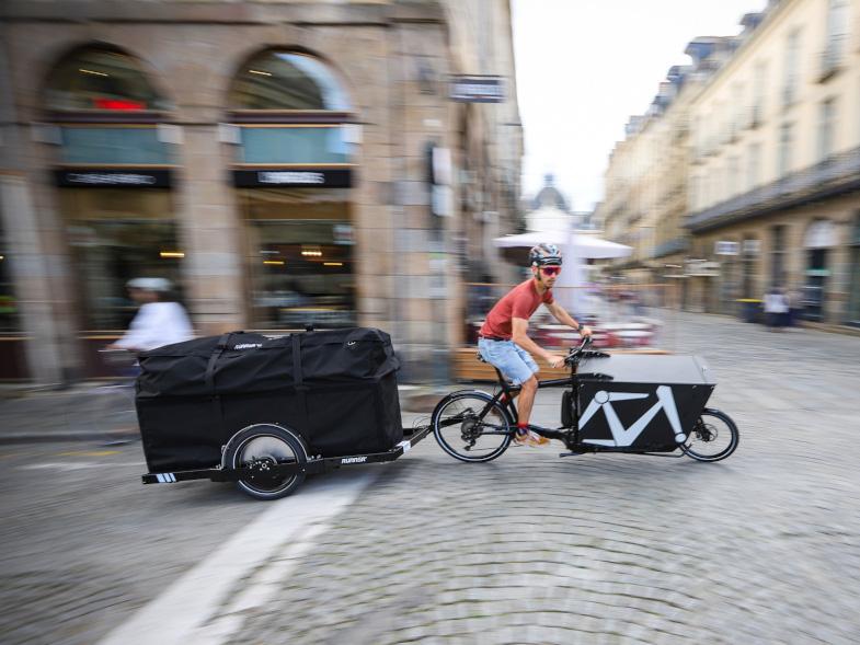 Bike courier on a Bullitt bike pulling a Runner bicycle trailer with a MegaBag on top of it, the courier is in a city center