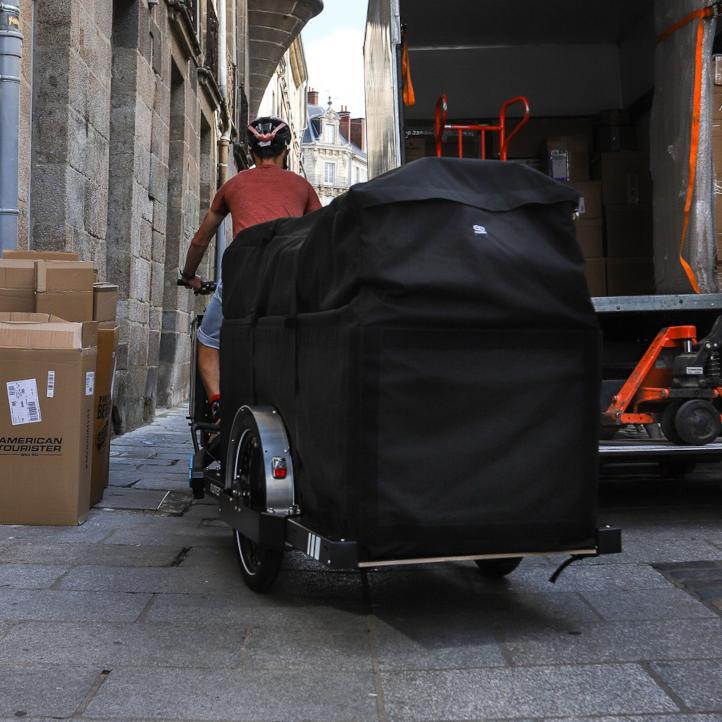 Bike courier pulling a trailer with a big black bag behind him passing next to a parked truck
