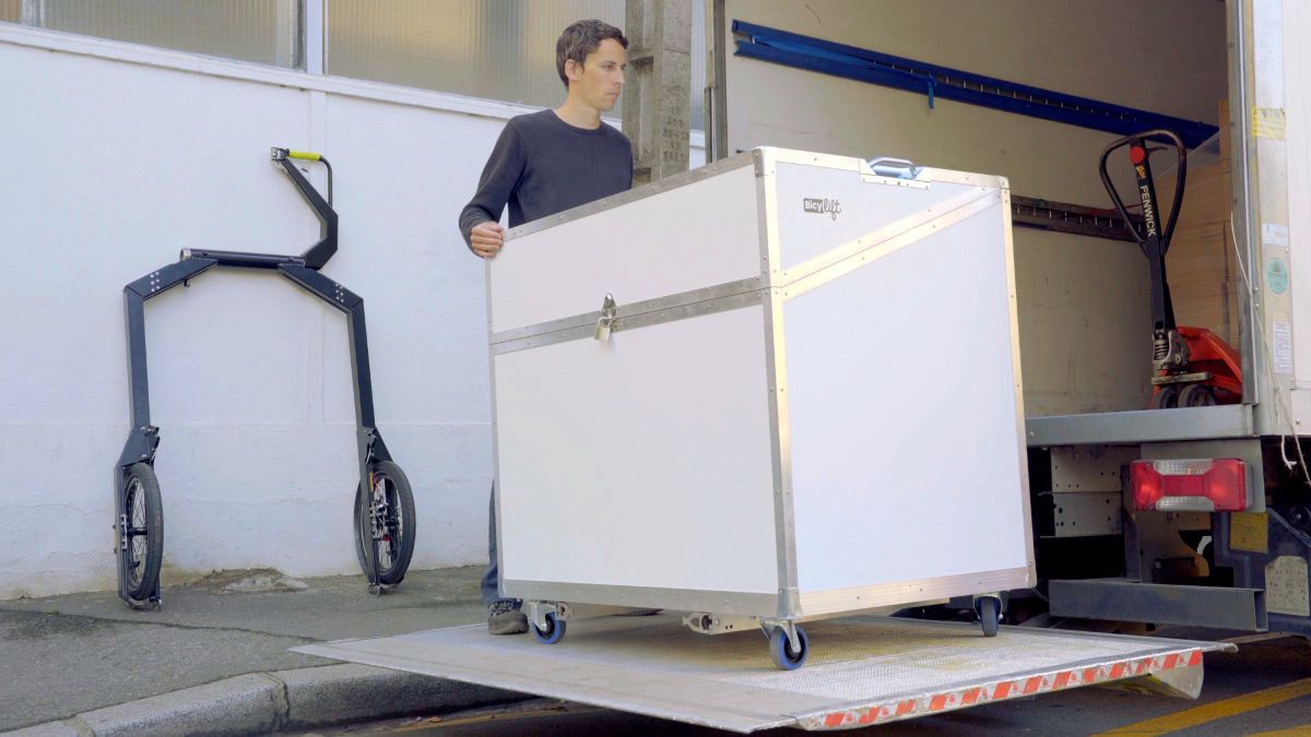 Someone with his hands on a white container on the elevator of a truck, a BicyLift trailer is resting against a white wall